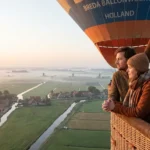 Een koppel staat samen in een luchtballon mand en kijkt uit over een mistig landschap met weilanden en waterwegen beneden.