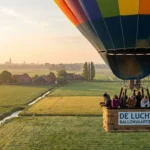 Kleurrijke heteluchtballon stijgt op boven Brabantse weilanden met passagiers in mand, boerderijen en stadshorizon zichtbaar.